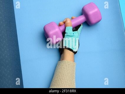Die weibliche Hand in einem blauen Sporthandschuh hält eine violette ein Kilogramm schwere Kurzhantel auf blauem Hintergrund. Stockfoto