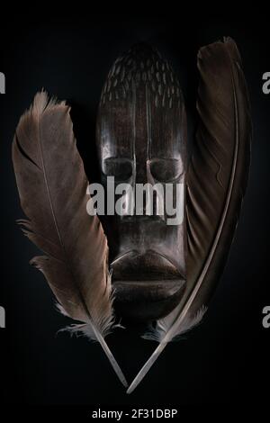 Holzskulptur des menschlichen Gesichts im afrikanischen Stil mit großen Federn um ihn herum. Holzschnitzerei aus dunklem Holz. Afrikanische Stammesmaske isoliert auf schwarzem Hintergrund. Kunst Stockfoto