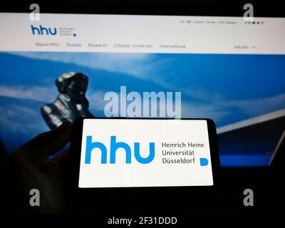 Person mit Mobiltelefon und Logo der Heinrich-Heine-Universität Düsseldorf (HHU) auf dem Bildschirm vor der Webseite. Konzentrieren Sie sich auf die Telefonanzeige. Stockfoto