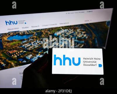 Person, die das Smartphone mit dem Logo der Heinrich-Heine-Universität Düsseldorf (HHU) auf dem Bildschirm vor der Webseite hält. Konzentrieren Sie sich auf die Anzeige des Mobiltelefons. Stockfoto