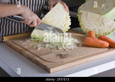 Auf einem Schneidebrett sind drei bunt gefärbte Karotten und ein riesiger Kopf von saftigem Kohl in zwei Hälften geschnitten, eine Frau schneidet den Kohl mit einem großen Messer. Stockfoto