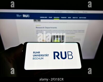 Person, die das Mobiltelefon mit dem Logo der deutschen Bildungseinrichtung Ruhr-Universität Bochum auf dem Bildschirm vor der Webseite hält. Konzentrieren Sie sich auf die Telefonanzeige. Stockfoto