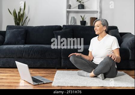 Lächelnde und aktive ältere Frau sitzt auf dem Boden in Lotushaltung, schaut Online-Yoga-Tutorial auf dem Laptop, ältere Frau Yoga zu Hause mit Video-Kurse, Online-Training, gesunde Lebensweise Stockfoto