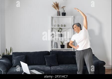 Aktive und sportliche ältere Frau, die sich vor dem Training zu Hause auswärmt, eine ältere Frau, die die Arme vor dem Laptop streckt, eine Dame mittleren Alters, die mit Video-Tutorial trainiert. Gesundes Lifestyle-Konzept Stockfoto