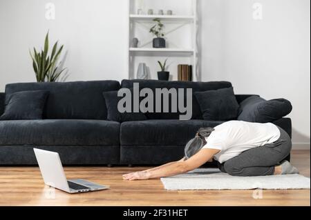 Ältere Frau praktiziert Yoga auf dem Boden vor dem Laptop-Computer, Online-Yoga-Tutorial, Ruhe nach aktiven Phasen, ältere Frauen Training mit Video-Klassen, genießt Online-Training Stockfoto