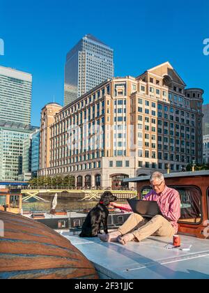 Geschäftsmann 50-55 Jahre, der mit seinem Spaniel-Hund draußen auf einem Bürokahnenboot arbeitet, Stadtbüros hinter Canary Wharf London E14 UK Stockfoto