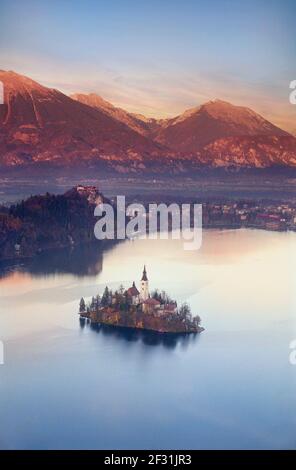 BLED-SEE INSEL SONNENUNTERGANG VERTIKALES PORTRÄT SLOWENIEN hoher Blick auf den See die Insel und die Kirche auf einem perfekten stillen Bleder See bei rotem Sonnenuntergang mit Herbstfarbe Julian Alps mit Sonnenuntergang hinter Slowenien Stockfoto