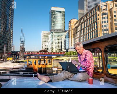 Home Working reifen Mann 50-55 draußen auf seinem Hausboot Barge, die Arbeit an seinem Computer kontrastiert mit der Umgebung von Bürogebäuden, Hotels und Cross-Rail-U-Bahn im Hintergrund. Canary Wharf London, Großbritannien. Telearbeit, auch Telearbeit genannt, Zukunft der Arbeit, Telearbeit, Telearbeit, Heimarbeit, Mobile Arbeit, Remote-Arbeit, Arbeit von überall und flexibler Arbeitsplatz, eine Arbeitsanordnung, bei der Mitarbeiter nicht pendeln oder an einen zentralen Arbeitsplatz, wie ein Bürogebäude, ein Lager oder ein Geschäft, reisen. Stockfoto