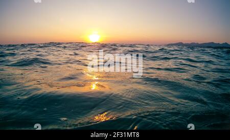 Orangefarbene Sonnenuntergangssonne, die über Meereswellen scheint, steht in Griechenland am klaren Himmel. Sommer ruhig Meerwasser Nahaufnahme am Abend Stockfoto