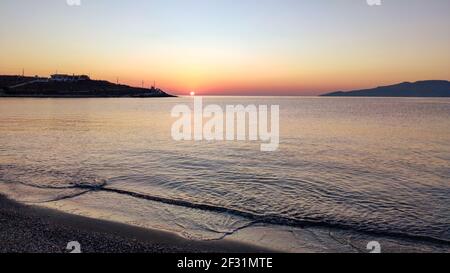 Orangefarbene Sonnenaufgangssonne, die am klaren Himmelshorizont an der Kiesstrand-Küste bei Athen in Griechenland scheint. Sommerurlaub ruhige Küste am Meer am Morgen Stockfoto