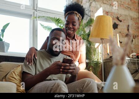 Fröhliche junge afrikanische Paar in Casualwear Blick auf Bildschirm Smartphone von lächelndem Mann gehalten, während er vor sitzt Kamera gegen Fenster Stockfoto