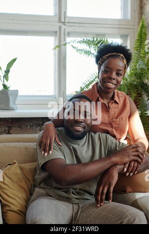 Glücklich junge afrikanische Paar in Casualwear Blick auf Sie mit Lächelt, während man vor der Kamera gegen das Fenster sitzt Grüne heimische Pflanzen Stockfoto