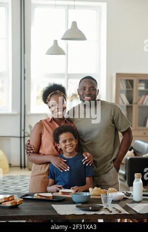 Fröhliche afrikanische Familie, bestehend aus Mutter, Vater und niedlichen kleinen Sohn stehen am Küchentisch vor der Kamera in häuslicher Umgebung Stockfoto
