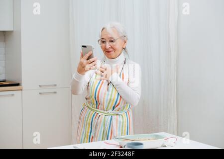 Freundliche Oma, die per Videoanruf in der Küche telefoniert und ihren Fortschritt teilt. Sie lächelt sanft und hält ihre Hand auf einer Brust. Stockfoto