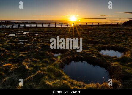 East Lothian, Schottland, Großbritannien, 14th. März 2021. UK Wetter: Sonnenuntergang im Aberlady Naturreservat. Die Sonne geht über das Feuchtgebiet in Aberlady Bay und die hölzerne Fußgängerbrücke unter und erleuchtet einen Neumond am Himmel Stockfoto