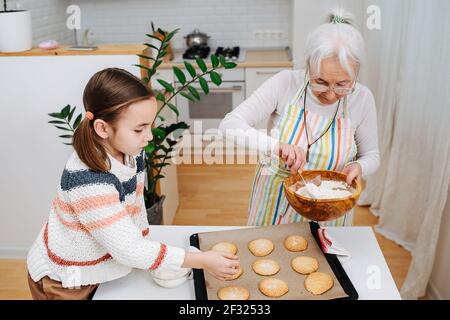 Scharf Oma streuen Zucker Pulver auf frischen Cookies neben ihrer Enkelin. Zeit zusammen mit dem Backen verbringen. Stockfoto