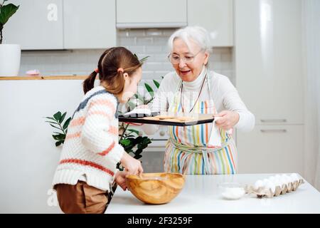 Virtuose Oma kochen mit ihrer Enkelin. Sie zeigt ihr frisch gebackene Kekse, während sie Eier hackt. Stockfoto