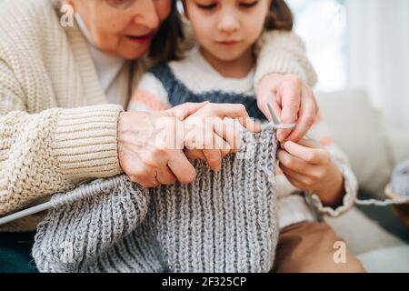Enthusiastische Oma, die mit ihrer Enkelin auf einer Couch sitzt, lehrt sie, wie man strickt, wobei sie die Hände über ihre hält. Nahaufnahme, Fokus auf Hände. Gesichter