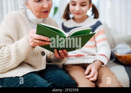 Gute Oma sitzt auf einer Couch mit ihrer Enkelin. Sie lesen zusammen ein Buch. Fokus auf das Objekt, Gesichter verschwommen. Stockfoto