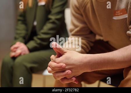 Zusammengekrallte Hände des jungen männlichen Patienten der Gruppe der geistigen Unterstützung Sitzen vor der Kamera unter anderen Menschen während psychologischer Sitzung Stockfoto