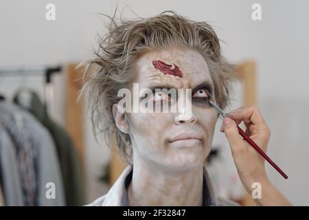 Hand der weiblichen Make-up-Künstler mit Pinsel Anwendung Zombie-Schmierfett Auf dem Unteraugen-Bereich des jungen Geschäftsmannes oder Schauspielers im Büro Oder Studio Stockfoto