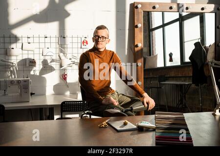 Junger seriöser männlicher Schneider mit Prothese statt linkem Bein Sitzen am Arbeitsplatz vor der Kamera in großen zeitgenössischen Werkstatt Stockfoto