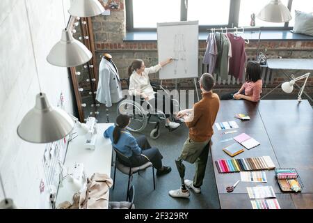 Junge kreative Designer im Rollstuhl zeigt auf Skizze auf Whiteboard Während der Präsentation ihrer Teil des neuen Projekts zu Kollegen Stockfoto