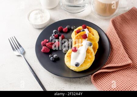 Quark Pfannkuchen auf einem schwarzen Teller mit Beeren, Sahne, Marmelade und Tasse Latte. Gesundes Frühstück. Hausgemachtes Essen Stockfoto