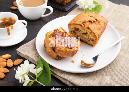 Hausgemachter Kuchen mit Rosinen, Mandeln, weichem Karamell und einer Tasse Kaffee auf schwarzem Betonboden und Leinentextilien. Seitenansicht, Nahaufnahme. Stockfoto