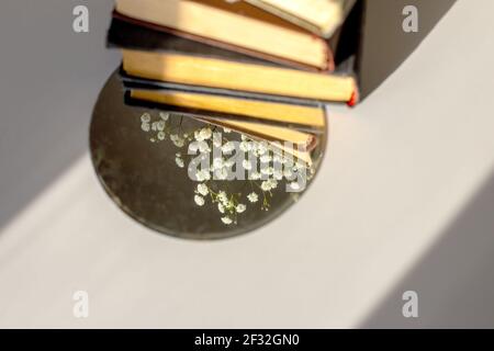 Drei alte Bücher liegen mit weißen Gypsophila-Blüten und einem Runder Spiegel auf dem Tisch im Sonnenlicht Stockfoto