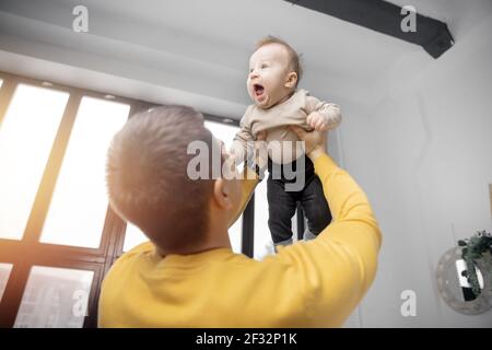 Glücklicher Vater spielt mit dem kleinen Sohn zu Hause vor dem Fenster, wirft sich auf, Junge lacht und lächelt. Stockfoto