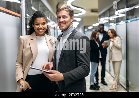 Glückliche multirassische Kollegen stehen in der Bürohalle und arbeiten zusammen an Unternehmensprojekt. Erfolgreiche Geschäftspartner blicken auf die Kamera, lächeln, Teamwork-Konzept Stockfoto