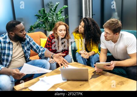 Konzentrierte multirassische Geschäftspartner diskutieren Unternehmensprojekt, reden über verschiedene Ideen. Junge Erwachsene Kollegen oder Studenten sitzen auf dem Sofa, mit Laptop, arbeiten oder studieren zusammen Stockfoto