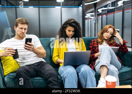 Seriöse Mischrennenkollegen sitzen auf dem Sofa im modernen Büro, suchen Ideen für Projekt, arbeiten zusammen. Fokussierte junge Erwachsene Mitarbeiter in stilvoller Kleidung mit Gadgets, Teamwork-Konzept Stockfoto