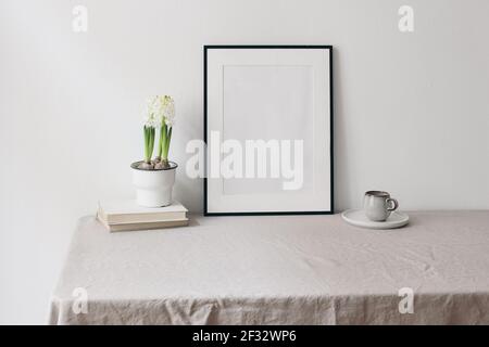 Ostern Frühling Stillleben. Blank schwarzer Bilderrahmen Mockup auf Leinentischtuch. Alte Bücher über Tasse Kaffee und Hite Hyazinthe in Blumentopf. Zu Hause Stockfoto