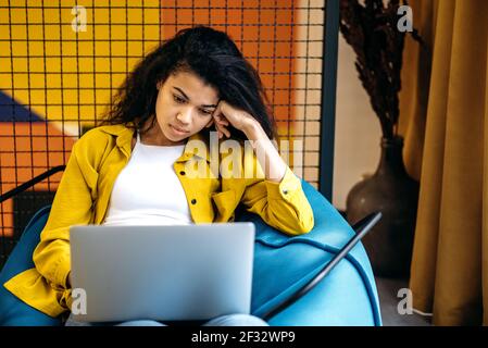 Müde junge Frau ist mit Laptop, sitzen im Stuhl zu Hause oder im Büro. afroamerikanische Mitarbeiterin oder Studentin wird beim Studium oder Arbeiten gelangweilt und fühlt sich erschöpft Stockfoto