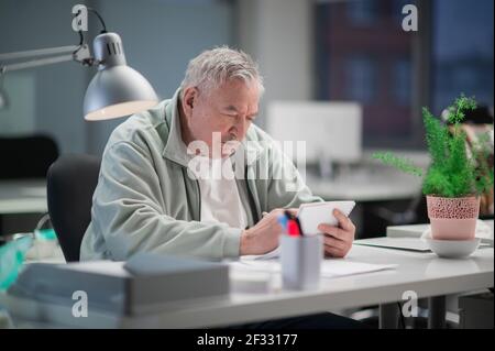 Ein älterer grauhaariger Büroangestellter an seinem Schreibtisch liest einen Text in einem Notizbuch. Ein großes, helles Büro mit Pflanzen und einem Computer im Hintergrund Stockfoto