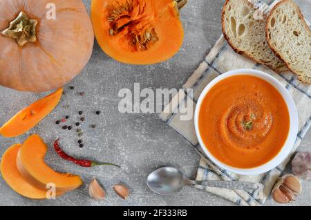 Pikante Kürbissuppe in einer weißen Schüssel neben frischem Kürbis, Brotscheiben und Gewürzen auf grauem Grund, flach auflegen. Gesunde Ernährung Konzept. Stockfoto