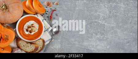 Frisch zubereitete pikante Kürbiscremesuppe mit Sauerrahm und Croutons in einer weißen Schüssel neben frischem Kürbis, Brotscheiben und Gewürzen auf einem grauen Backg Stockfoto