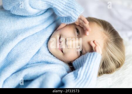 Niedliche kleine kaukasische Mädchen sieben Jahre alt mit blonden Haaren liegen auf dem Bett zu Hause im Schlafzimmer. Kind trägt modisch gemütlich Strickpullover blau Farbe Stockfoto