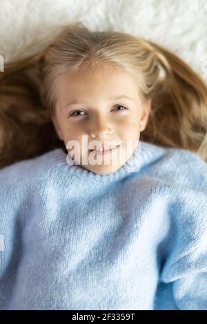 Niedliche kleine kaukasische Mädchen sieben Jahre alt mit blonden Haaren liegen auf dem Bett zu Hause im Schlafzimmer. Kind trägt modisch gemütlich Strickpullover blau Farbe Stockfoto