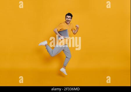 In voller Länge Foto von einem fröhlichen lustigen kaukasischen smart jungen Mann in Brillen, stilvoll gekleidet, hält Laptop, lächelnd und springen hoch läuft auf isolierten orangen Hintergrund Stockfoto