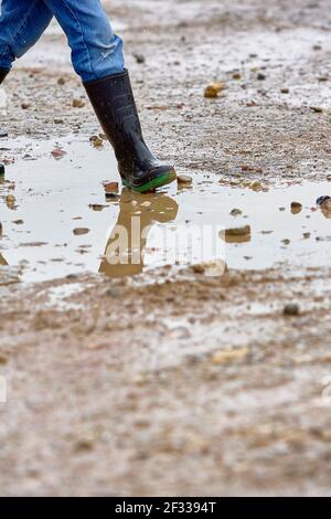 Mädchen mit Gummistiefeln, die in einer schlammigen Pfütze auf nassem Kies stehen und das Wasser reflektiert ihre Stiefel. Stockfoto