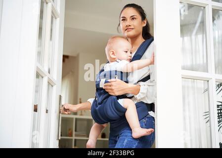 Hübsche junge Frau mit gemischter Rasse, die die Eingangstür öffnete und das Haus verließ Mit Baby Junge in Träger Stockfoto