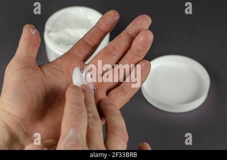 Hand reibt kosmetische Creme in die Hand. Ein Mann mittleren Alters kümmert sich um die Haut seiner Hände. Ein Behälter mit Sahne und einem Deckel im Hintergrund. Stockfoto