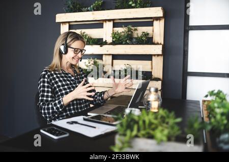Die hübsche, junge blonde Geschäftsfrau mit schwarz-weiß kariertem Hemd sitzt in einem nachhaltigen, ökologischen Büro mit Headset und hat ein Web Stockfoto