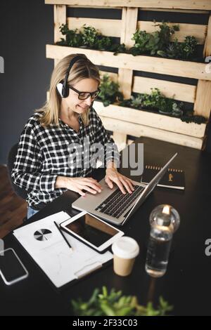 Die hübsche, junge blonde Geschäftsfrau mit schwarz-weiß kariertem Hemd sitzt in einem nachhaltigen, ökologischen Büro mit Headset und hat ein Web Stockfoto