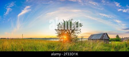 Panoramablick auf Sonnenuntergang über alte Holzhütte und einsam Baum auf dem Land Stockfoto