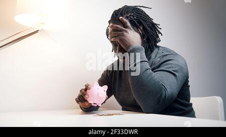 Leeres Sparschwein. Trauriger afroamerikanischer Mann, der eine Münze ins Innere steckt. Geringe Einsparungen. Hochwertige Fotos Stockfoto