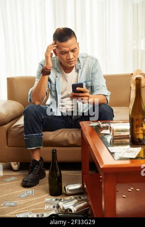 Betrunkener junger asiatischer Mann mit Kopfschmerzen, der auf dem Sofa sitzt Zuhause mit leeren Flaschen und Dosen herum und Text lesen Nachricht auf dem Smartphone Stockfoto